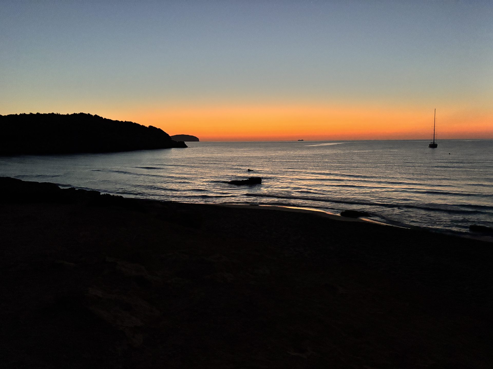 Cala Nova beach in Ibiza with an orange sky, calm sea and an anchored sailboat